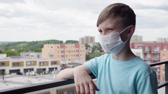 A Young Boy in a Face Mask Stands on a Balcony and Looks Over the Cityscape Below - Closeup