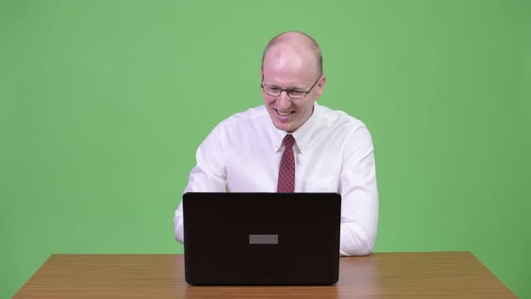 Happy Mature Bald Businessman Using Laptop and Getting Good News Against Wooden Table