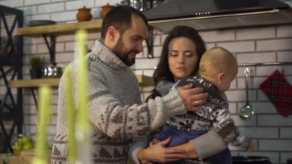 Bearded Father in Warm Sweater Playing with Baby Little Son Who Is in Mother Arms in the Kitchen