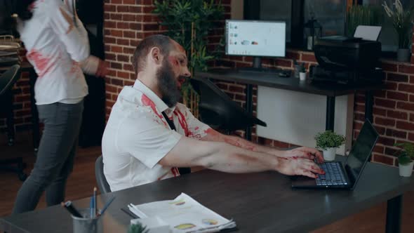 Scary Office Zombie with Bloody Wounds in Workspace Working on Modern Laptop