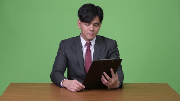 Young Handsome Asian Businessman Working with Clipboard Against Green Background