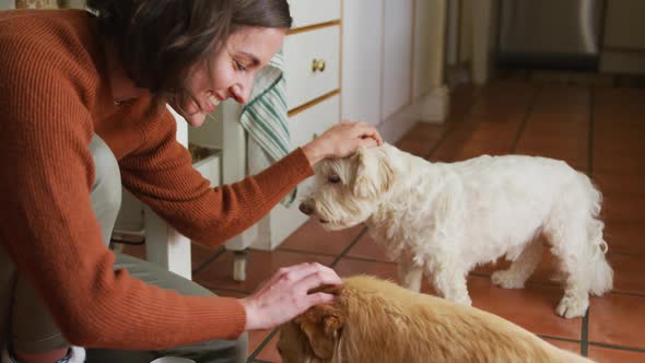 Smiling caucasian woman stroking her pet dogs eating from bowl in kitchen at home