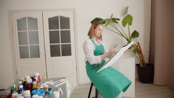 Young Woman Draws on Canvas with Brush Sitting Chair in Living Room There are Jars of Paint on Table