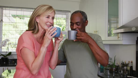 Happy diverse senior couple drinking coffee and talking standing in kitchen