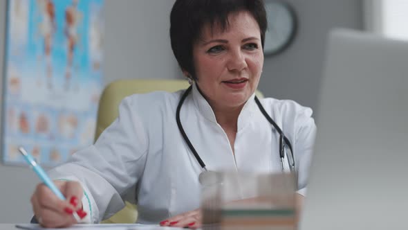 Middle Aged Senior Head Doctor Sitting at Workplace Talking to Patient Making Video Call on Laptop