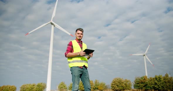 The Engineer Work Next to the Windmill Turbines with the Tablet in His Hand