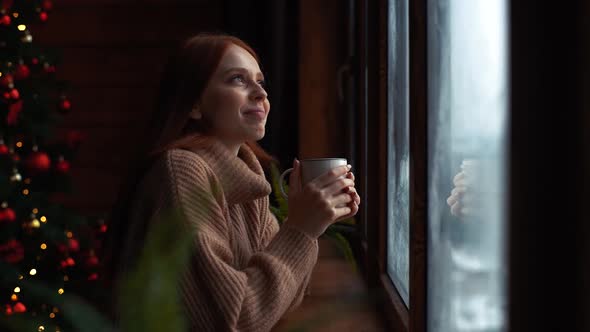 Side View of Happy Young Redhead Woman with Cup Standing By Window at Home at Christmas Time