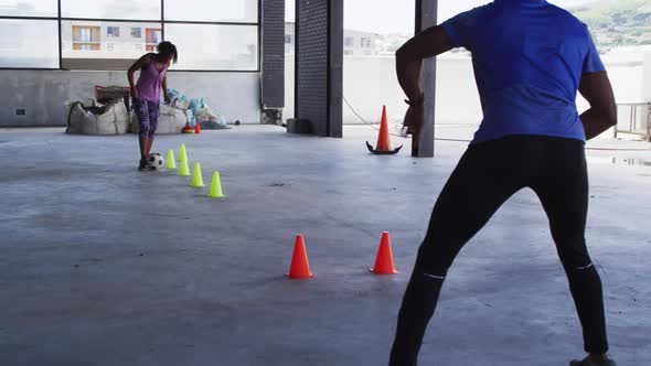 African american woman doing slalom with a football in urban building with man cheering her