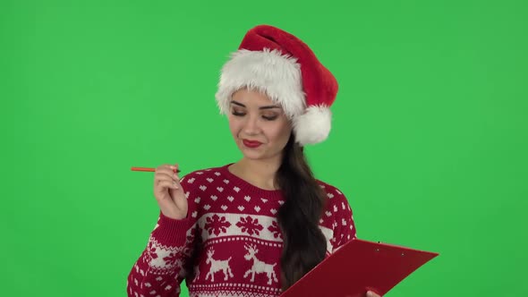 Portrait of Sweety Girl in Santa Claus Hat Fills Papers in Red Folder with Pensil. Green Screen