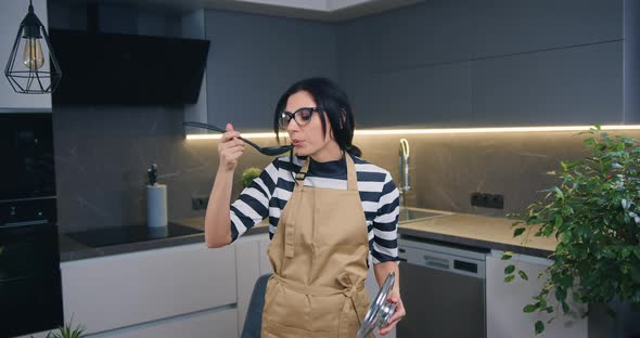 Young Woman in Apron Prepared Soup and Tasting it in Modern kitchen