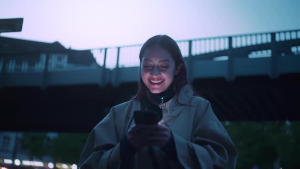 Young woman surrounded with evening city lights she is looking at her phone and laughs