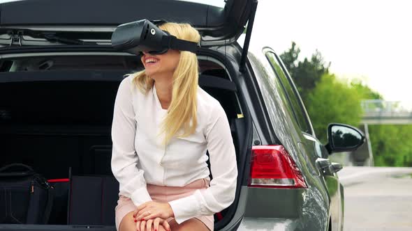 Young Attractive Blond Woman Sits in the Trunk and Uses Virtual Reality Glasses