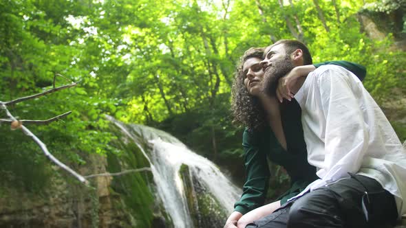 Young Couple - Man with Beard and Woman with Curly Hair Is Hugging and Kissing By Sitting on Stone