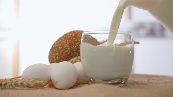 Pouring Fresh Milk Into A Glass On A Rustic Straw Table, Close Up Of White Chicken Eggs With Wheat