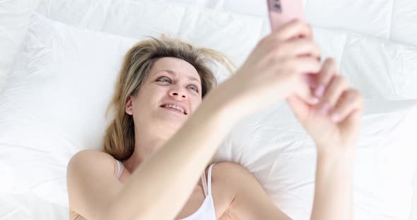 Attractive Young Woman Smiling and Looking at Smartphone Camera While Lying on Bed at Home