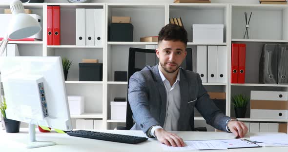 Businessman in Gray Jacket Sitting at Table in White Office and Offering Documents