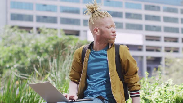 Thoughtful albino african american man with dreadlocks sitting in park using laptop