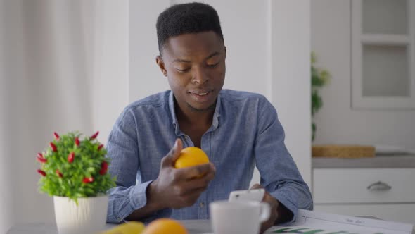 Front View of Happy Man Sitting at Kitchen Table Messaging on Smartphone Juggling Orange in Hand