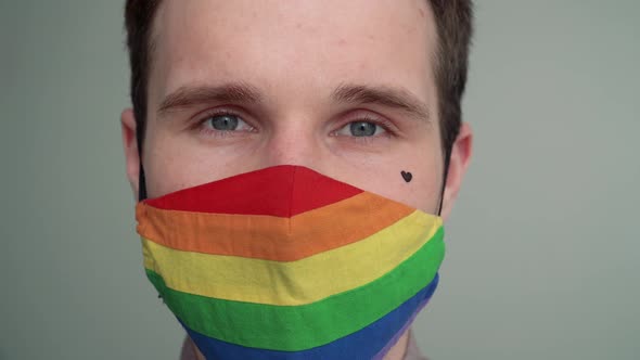 Close-up portrait of a young man with a black heart on his cheek taking off a rainbow face mask