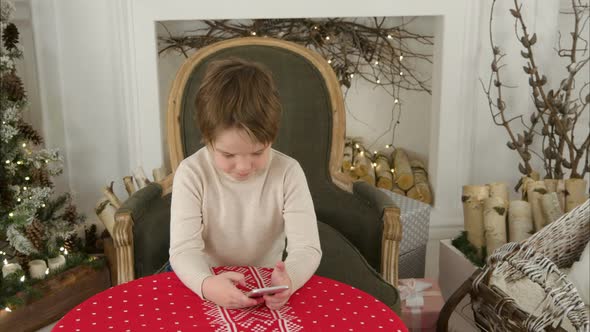 Boy Playing Games on His Phone Sitting at the Table Near the Christmas Tree