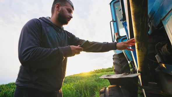 Man in Field with Phone in His Hands Stands By His Tractor and Knocks on Wheel Checking Condition of