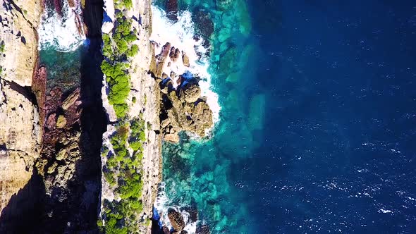 Drone looking straight down while flying over long narrow rock structure and sea side cliffs surroun