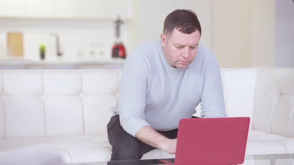 Concentrated Caucasian Man Messaging Online on Laptop Sitting on Couch at Home