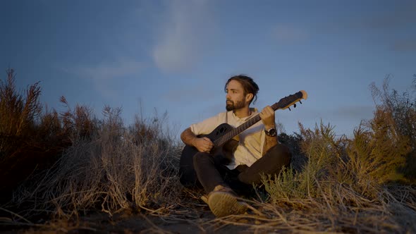 A Young Man Sitting on a Grassy Meadow and Playing Guitar at Sunset  Slowmotion Pull Back