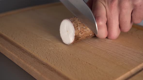 A man cuts with a knife the yucca root on a wooden kitchen board on a gray table