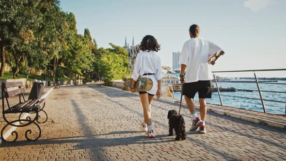 Back View of Couple with Skateboard Walking Their Dog in Evening on Embankment Slow Motion