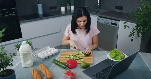 Brunette in Modern Cuisine and Cutting Cucumber for Salad Simultaneously Choosing Program on Laptop