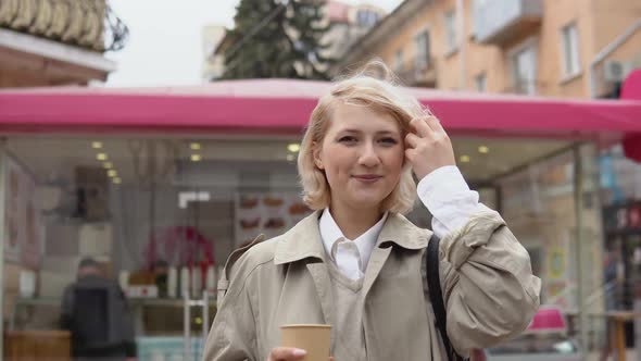 Young Blonde Business Woman in a Beige Trench Coat and Vest a White Blouse Drinks Takeaway Coffee in