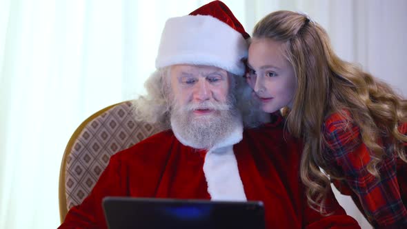 Portrait of Old Santa Sitting with Tablet As Cheerful Little Girl Whispering on Ear