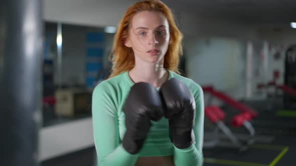 Perspiring Satisfied Redhead Sportswoman Hitting Boxing Gloves Looking at Camera with Blurred Woman