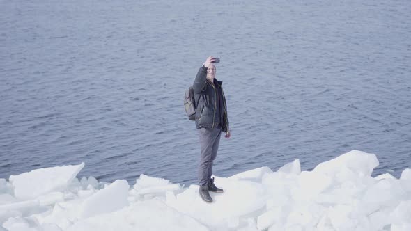 The Man in a Warm Coat and Hat Standing on the Edge of Glacier Taking Selfie