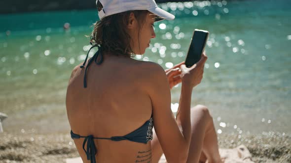 Rear View Tanned Woman Tourist in Bikini Sits on Sea Beach Close to Water Unlocks Her Mobile Phone