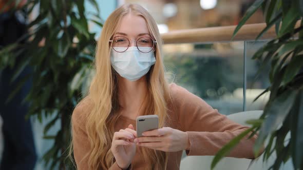 Excited Blonde Caucasian Woman 20s Woman Wears Medical Mask Indoor Sitting on Sofa Use Mobile Phone