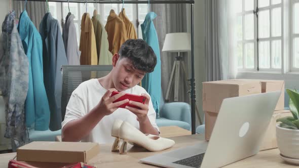 Asian Man Online Seller Taking Photos With Mobile Phone While Using Computer For Selling Clothes