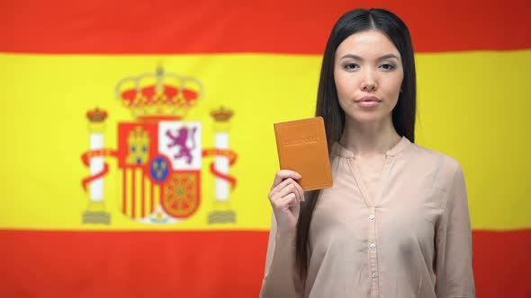 Serious Asian Woman Showing Passport Against Spanish Flag Background, Migration