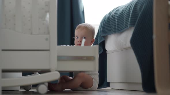 Cute Small Child Sitting on the Floor Explores Shelf Under the Crib 1 Year Old Baby Boy Playing in