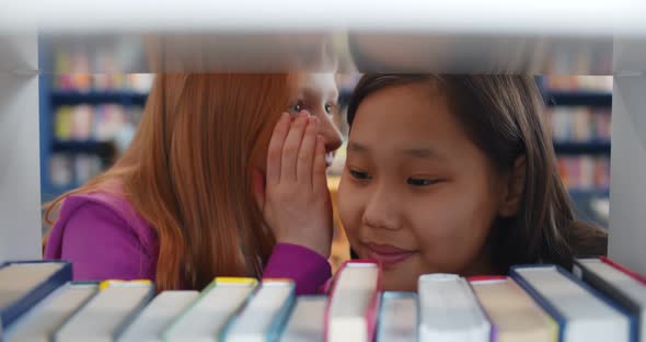 Teen Girl Students in Library Whispering Secrets Standing Behind Bookshelf