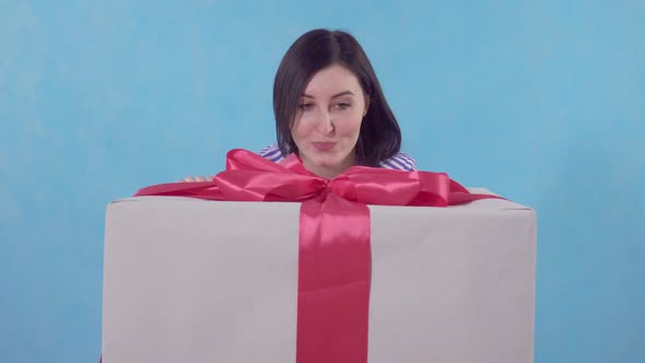 Funny Young Woman Looks Out From Behind a Large Packaged Gift on a Blue Background