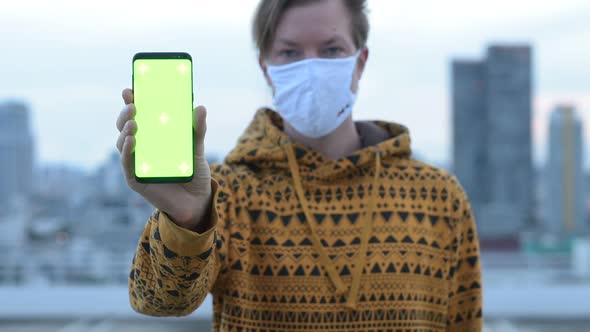 Face of Young Man with Mask Showing Phone Against View of the City