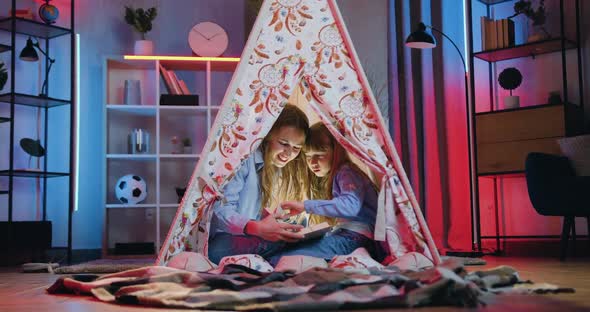 friendly two different ages sisters sitting in wigwam at night and reading interesting book