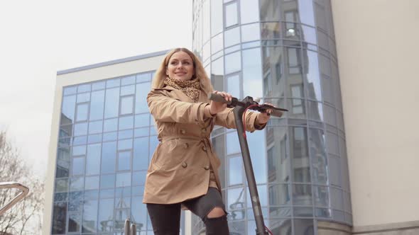 Young Stylish Blonde Girl in a Beige Trench Coat and Jeans Stands with an Electric Scooter on the