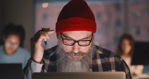 Portrait of Senior Hipster Bearded Man Holding Pencil Working on Laptop in Office