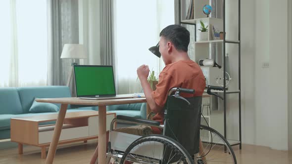 Young Asian Boy Sitting In A Wheelchair While Using Laptop With Mock Up Green Screen And Celebrating