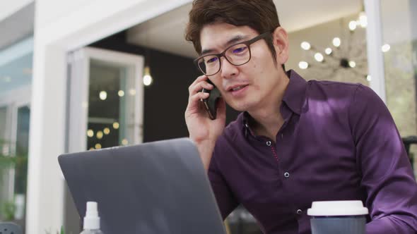 Asian man talking on smartphone and using laptop while sitting at a cafe