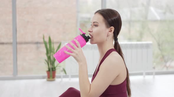 a Young Woman in a Burgundy Tracksuit Drinks Water on a Mat in a Gym