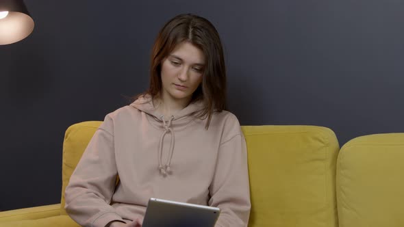 Young Lady Holds Tablet Communicating on Social Networks on Sofa in Cozy Room